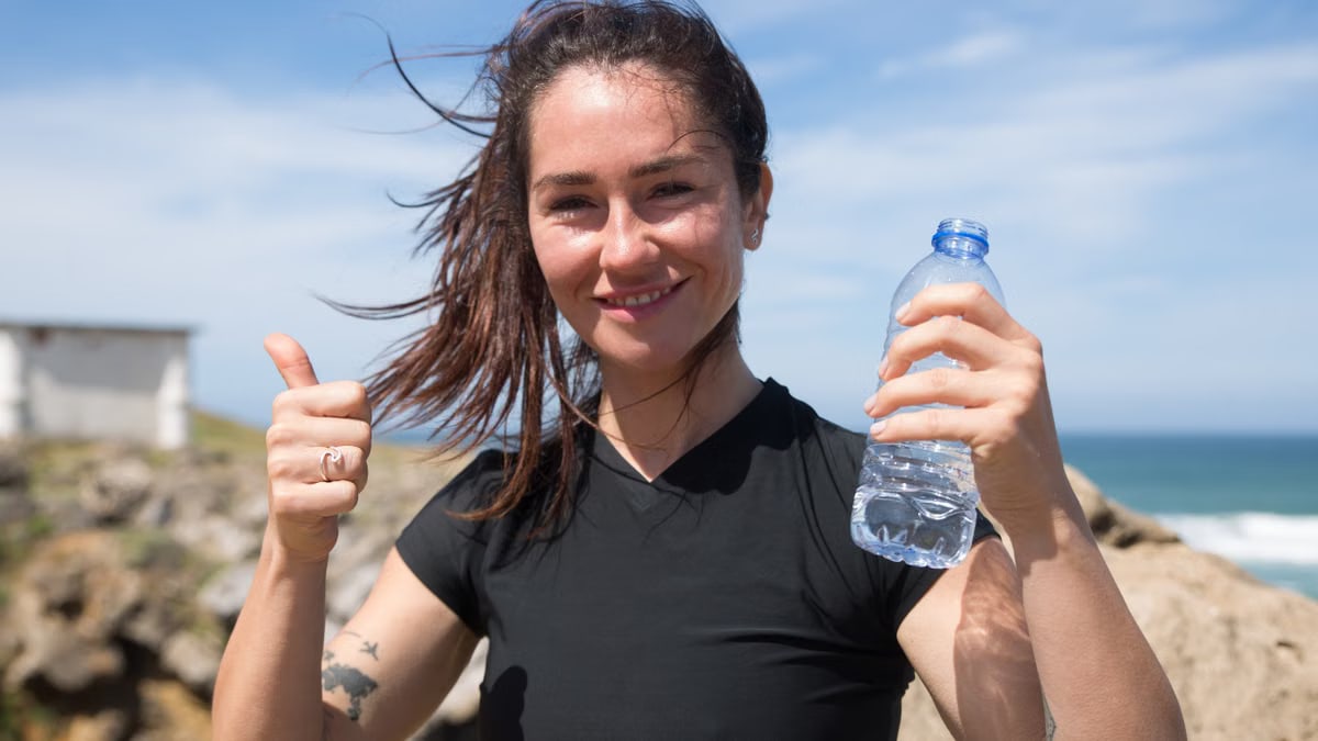 Happy woman giving thumbs up with water bottle on a sunny beach day in Portugal.